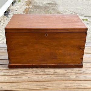 May include: A rectangular wooden chest with a hinged lid. The chest is made of a warm-toned wood, possibly mahogany or walnut, and features a keyhole on the front. The chest is constructed with dovetail joints and sits on a low base.