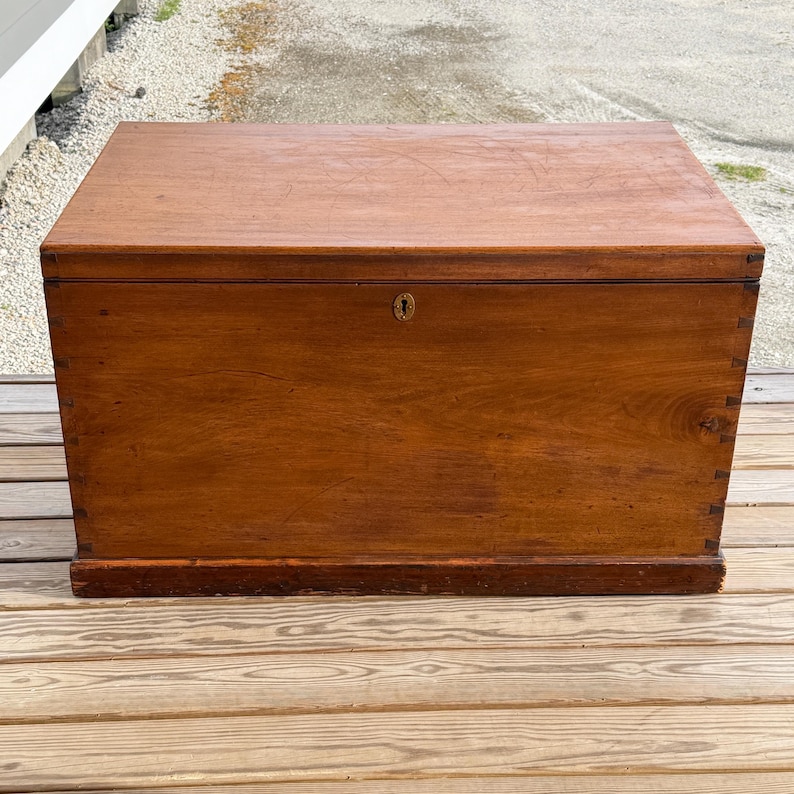 May include: A rectangular wooden chest with a hinged lid. The chest is made of a warm-toned wood, possibly mahogany or walnut, and features a keyhole on the front. The chest is constructed with dovetail joints and sits on a low base.