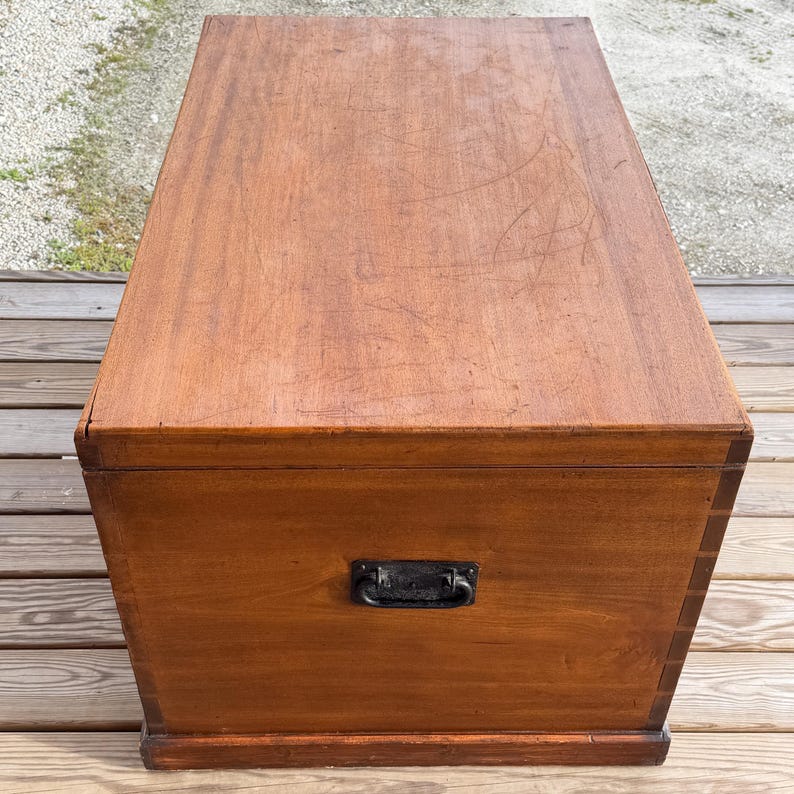 May include: A rectangular wooden chest with a dark brown finish. The chest has a black metal handle on the front and is sitting on a wooden deck. The top of the chest shows signs of wear.