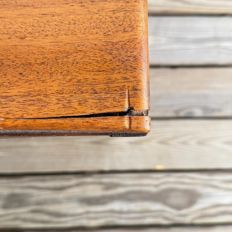 May include: Close-up of a wooden object with a warm brown finish. The wood grain is visible, and there is a crack along the edge. The object appears to be part of a larger piece of furniture.