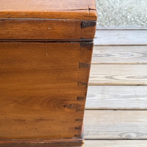 May include: Close-up of a wooden chest with dovetail joints. The chest is made of a warm-toned wood, possibly walnut or mahogany, with visible wood grain. The corners are reinforced with darker wood pieces, adding to its antique appearance.