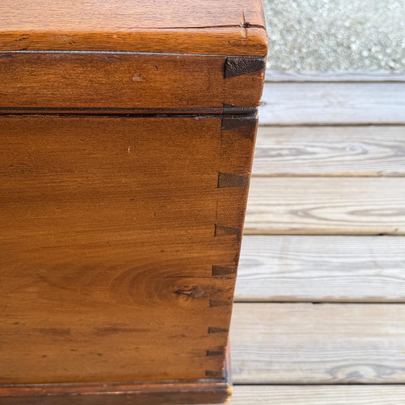 May include: Close-up of a wooden chest with dovetail joints. The chest is made of a warm-toned wood, possibly walnut or mahogany, with visible wood grain. The corners are reinforced with darker wood pieces, adding to its antique appearance.
