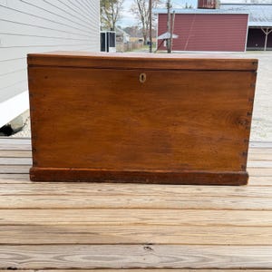 May include: A large, antique wooden chest with a warm brown finish. The chest features a rectangular shape with a slightly curved top and a brass keyhole. The wood grain is visible, and the chest sits on a low base.