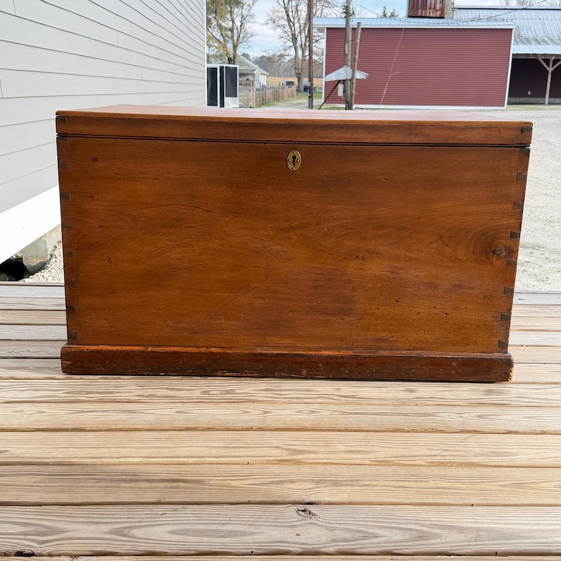 May include: A large, antique wooden chest with a warm brown finish. The chest features a rectangular shape with a slightly curved top and a brass keyhole. The wood grain is visible, and the chest sits on a low base.