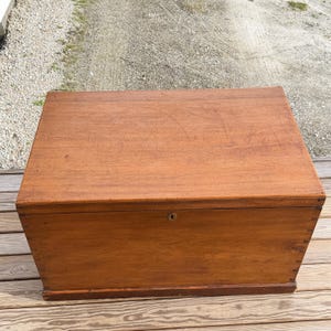 May include: A rectangular wooden chest with a warm brown finish. The chest has a simple design with a keyhole and is likely used for storage. The wood grain is visible on the surface, and the chest appears to be well-constructed.