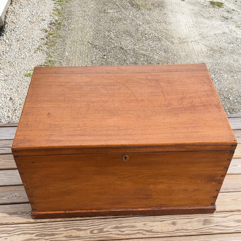May include: A rectangular wooden chest with a warm brown finish. The chest has a simple design with a keyhole and is likely used for storage. The wood grain is visible on the surface, and the chest appears to be well-constructed.