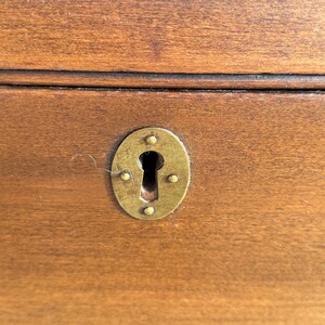 May include: Close-up of a wooden surface with a brass-colored oval keyhole plate. The keyhole plate is secured with four small rivets. The wood has a warm, brown tone with visible grain.