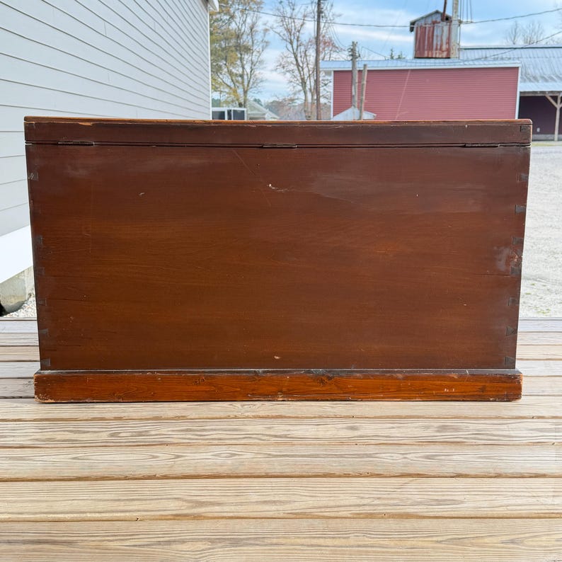 May include: A large, rectangular wooden chest with a dark brown finish. The chest has a hinged lid and is constructed with visible dovetail joints. The base of the chest is a lighter shade of brown, and it sits on a wooden deck.