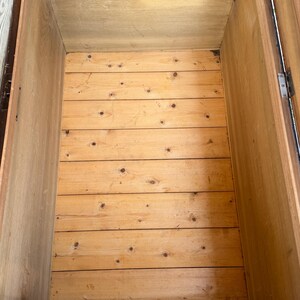 May include: An empty wooden chest interior with a light brown finish. The base is constructed of horizontal planks, and the sides are vertical. The chest appears to be aged, with visible wood grain and knots.