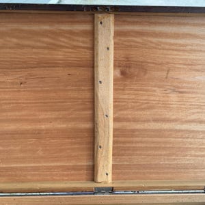 May include: Close-up of the interior of a wooden chest. The chest's interior is a warm, reddish-brown hue, with visible wood grain. A vertical wooden support beam is attached with small metal screws. The chest's edges are framed with a darker material.