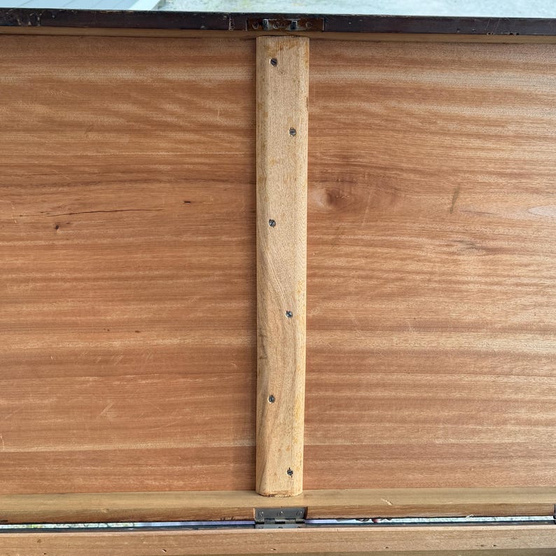 May include: Close-up of the interior of a wooden chest. The chest's interior is a warm, reddish-brown hue, with visible wood grain. A vertical wooden support beam is attached with small metal screws. The chest's edges are framed with a darker material.