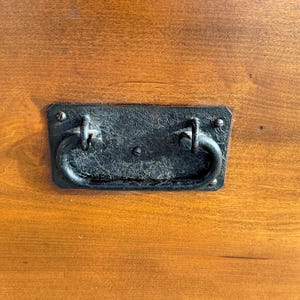 May include: Close-up of a rectangular, black metal handle with a curved, ring-shaped grip. The handle is attached to a wooden surface with visible wood grain. The wood has a warm, reddish-brown color.