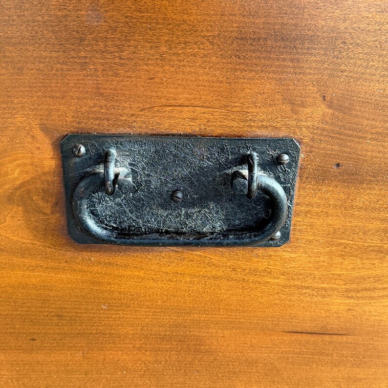 May include: Close-up of a rectangular, black metal handle with a curved, ring-shaped grip. The handle is attached to a wooden surface with visible wood grain. The wood has a warm, reddish-brown color.