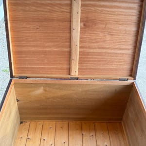 May include: An open wooden chest with a hinged lid. The interior and exterior are made of wood, with a natural finish. The lid is propped open, revealing the chest's interior. The chest appears to be vintage or antique, with visible wood grain.