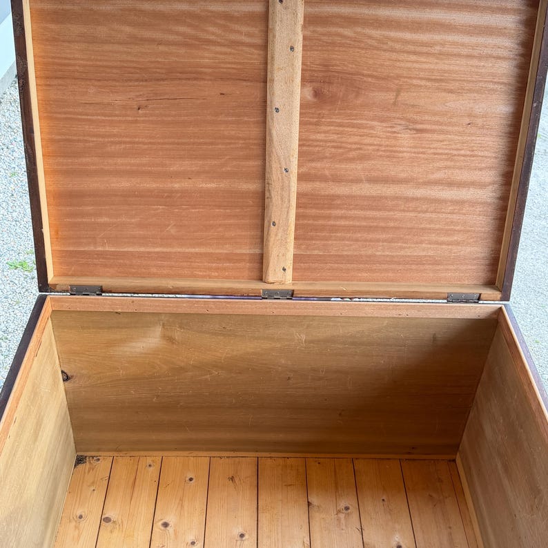 May include: An open wooden chest with a hinged lid. The interior and exterior are made of wood, with a natural finish. The lid is propped open, revealing the chest's interior. The chest appears to be vintage or antique, with visible wood grain.