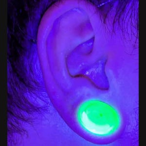 May include: Close-up of an ear with a large, glowing green plug. The ear is illuminated with a purple light, highlighting the skin and hair around it. The plug is circular and appears to be made of a translucent material.