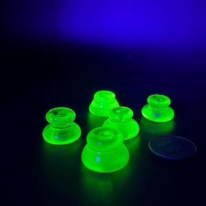 May include: A collection of six glowing green glass objects, possibly bottle stoppers, are arranged on a dark surface. The objects have a rounded base and a flared top. A coin is next to the objects.
