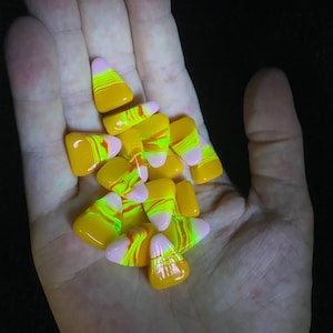 May include: A handful of candy corn candies. Each piece features a yellow base, a neon green and orange swirl, and a pink tip. The candies are illuminated against a dark background.