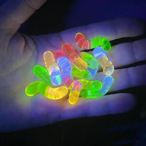 May include: A collection of glow-in-the-dark gummy worms in various colors, including green, yellow, orange, pink, and blue. The gummy worms are arranged in a pile on a person's hand.