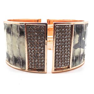 Peut inclure: Un large bracelet manchette avec une finition or rose. Le bracelet présente un motif texturé gris et blanc sur la bande principale, avec des sections rectangulaires de cristaux étincelants. Le bracelet est ouvert au centre.