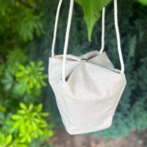 Peut inclure: Un sac en tissu beige clair avec des anses en corde blanche est suspendu &agrave; une branche d'arbre. Le sac a une forme carr&eacute;e avec un dessus pli&eacute; et un int&eacute;rieur visible. Le fond pr&eacute;sente un feuillage vert.