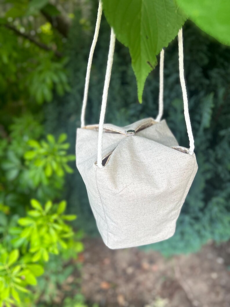 Peut inclure: Un sac en tissu beige clair avec des anses en corde blanche est suspendu &agrave; une branche d'arbre. Le sac a une forme carr&eacute;e avec un dessus pli&eacute; et un int&eacute;rieur visible. Le fond pr&eacute;sente un feuillage vert.