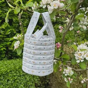 Op de afbeelding: Een kleine, gewatteerde stoffen draagtas met een bloemenpatroon in grijstinten, geel en blauw. De tas heeft twee handvatten en een trekkoordsluiting. De tas hangt aan een tak met witte en roze bloemen.
