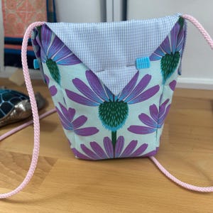 Peut inclure: Un petit sac carr&eacute; avec un fond bleu clair et un grand motif de fleurs violettes. Le rabat est en vichy bleu clair et blanc. Il a une bandouli&egrave;re en corde rose et des fermetures bleues.