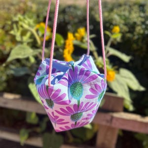 Peut inclure: Un sac en tissu suspendu avec un motif floral de fleurs violettes et blanches avec des centres verts. Le sac est suspendu par des poign&eacute;es en corde rose. L'arri&egrave;re-plan est flou, montrant de la verdure et des fleurs jaunes.