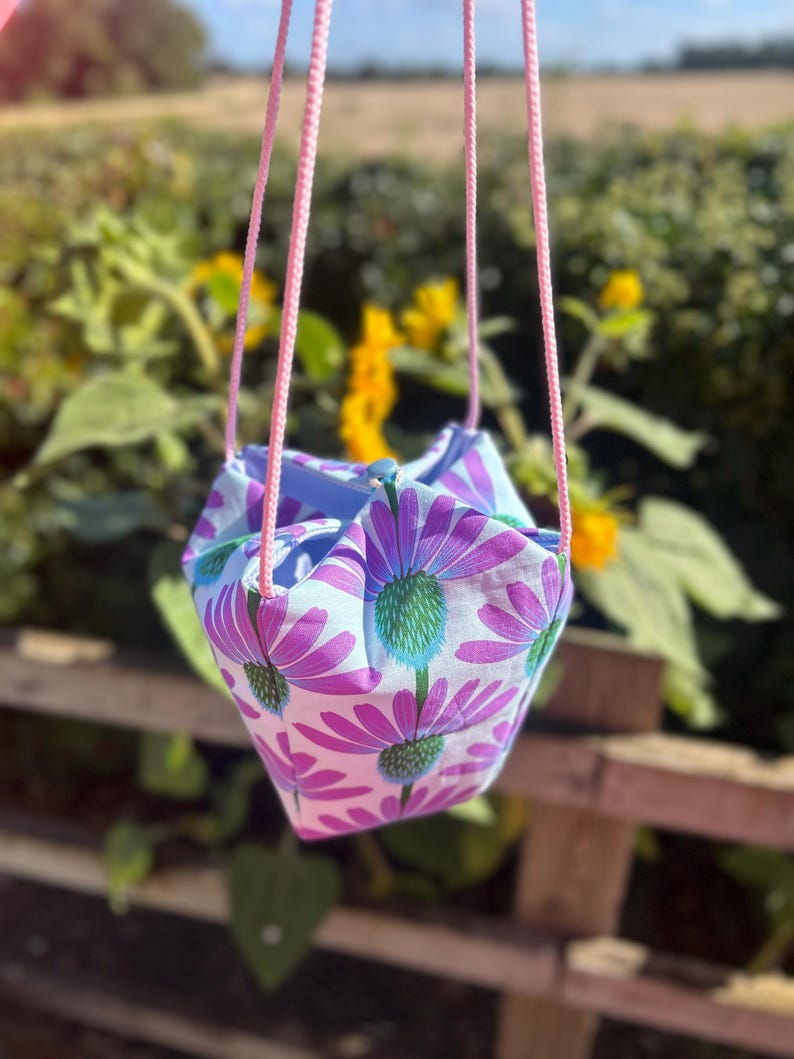 Peut inclure: Un sac en tissu suspendu avec un motif floral de fleurs violettes et blanches avec des centres verts. Le sac est suspendu par des poign&eacute;es en corde rose. L'arri&egrave;re-plan est flou, montrant de la verdure et des fleurs jaunes.