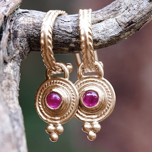 May include: A pair of gold-colored earrings with a circular design, each featuring a vibrant red gemstone center. The earrings are suspended from a textured, twisted loop and are displayed on a natural wooden branch.
