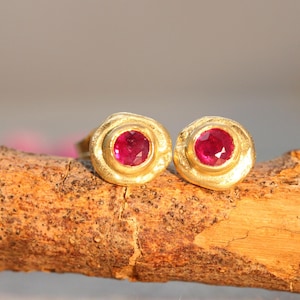 May include: A pair of gold stud earrings, each with a bright red gemstone. The earrings have a textured, organic design. The jewellery is displayed on a brown, textured surface.