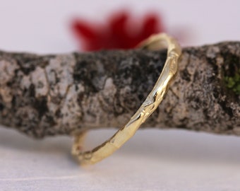 May include: A thin gold band ring with a textured surface, resembling a tree branch. The ring is resting on a piece of bark.