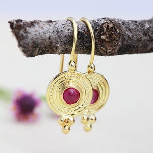 May include: Gold hoop earrings with a red gemstone in the center of a circular design. The earrings have a dangling charm with three small beads.