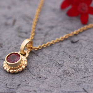 May include: A delicate gold chain necklace with a small red gemstone pendant. The pendant is surrounded by a beaded halo.