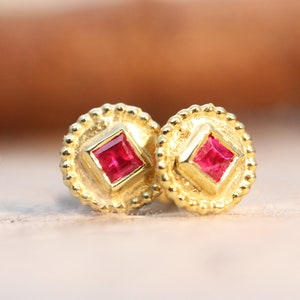 May include: A pair of gold earrings with a small square ruby gemstone set in each earring. The earrings have a textured, circular design.