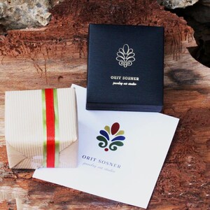 Op de afbeelding: Een klein, bruin cadeaudoosje met een rood en groen lint. Het doosje staat op een houten oppervlak. Een witte kaart met de tekst "ORIT SOSHER jewelry art studio" en een kleurrijk logo is op de achtergrond.