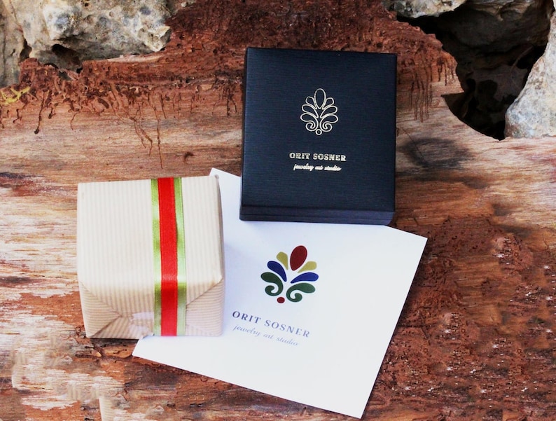 Op de afbeelding: Een klein, bruin cadeaudoosje met een rood en groen lint. Het doosje staat op een houten oppervlak. Een witte kaart met de tekst "ORIT SOSHER jewelry art studio" en een kleurrijk logo is op de achtergrond.