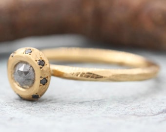 Op de afbeelding: Een gouden ring met een getextureerde, gehamerde band met een centrale ronde grijze steen omringd door een zetting met zes kleine zwarte steentjes. Het metaal heeft een matte afwerking en een ronde zetting die de stenen vasthoudt.