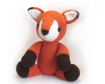 May include: A crocheted orange fox plush toy with brown accents on the ears and feet. The fox is sitting with its legs crossed.