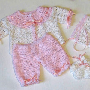 May include: A crocheted baby outfit in pink and white with a matching hat and booties. The outfit features a cardigan with a pink ribbon trim, pants with a pink ribbon trim, a hat with a pink ribbon trim, and booties with a pink ribbon trim.