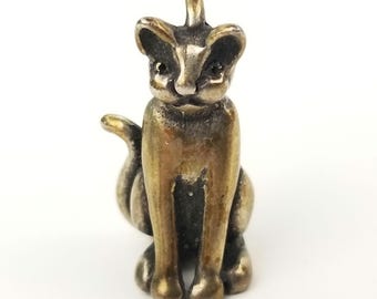 May include: A small, metallic cat figurine with a brass-toned finish and dark, recessed detailing. The cat is depicted in a seated position with its tail curled to the side and a small loop at the top of its head, suggesting use as a pendant or charm.