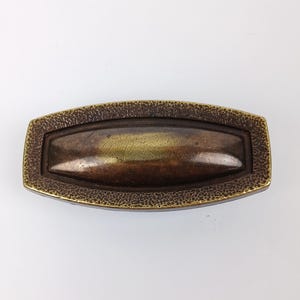 May include: An elongated, oval-shaped cabinet pull with a dark, aged bronze finish. The central section is smooth and slightly raised, while the surrounding border features a textured, hammered surface with golden highlights along the edges.