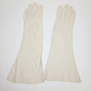 May include: A pair of long, off-white gloves reaching past the wrist. These gloves feature a smooth, matte finish with vertical stitched detailing along the back of the hand and fingers, extending into a flared, wide-opening cuff at the forearm.