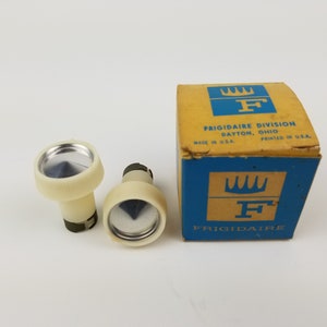 Może przedstawiać: Dwa białe plastikowe pokrętła z srebrnymi metalowymi wkładkami i pudełko z napisem "FRIGIDAIRE DIVISION DAYTON, OHIO" z niebiesko-żółtym logo.