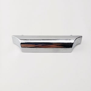 May include: A polished chrome cabinet pull with a curved design. The hardware features a rectangular body with flared ends, reflecting light and creating a sleek, modern aesthetic. The pull is designed for easy gripping and opening of drawers or cabinets.