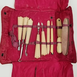 Può includere: Un astuccio arrotolabile in velluto rosso con un set di strumenti per la manicure vintage. Gli strumenti includono una lima per unghie, uno spingicuticole, una pinzetta e un tagliaunghie. Gli strumenti sono realizzati in avorio e metallo.