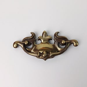 Puede incluir: Tirador de cajón de latón antiguo con un diseño decorativo de flor de lis. El herraje presenta un asa curvada con detalles ornamentados y un acabado cálido y envejecido. Perfecto para proyectos de restauración de muebles antiguos.