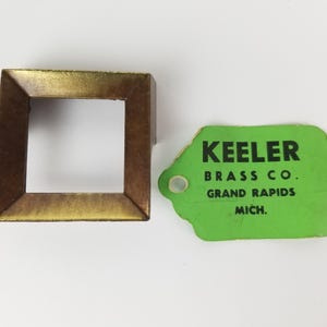 May include: A gold-coloured metal frame with a square opening. A green tag with the text "KEELER BRASS CO. GRAND RAPIDS MICH." is next to the frame.