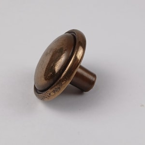 May include: A bronze cabinet knob with a rounded face and a short, cylindrical stem. The knob has a slightly distressed finish, giving it a vintage appearance. The design is simple and classic, suitable for various furniture styles.